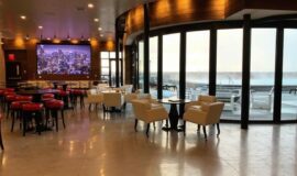 Infinity Room with large TV screen, tables and chairs, with floor to ceiling windows and views of rooftop swimming pool.