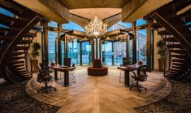 Grand entrance hall with curved dual staircases, wood and stone floors, and glowing glass chandelier.