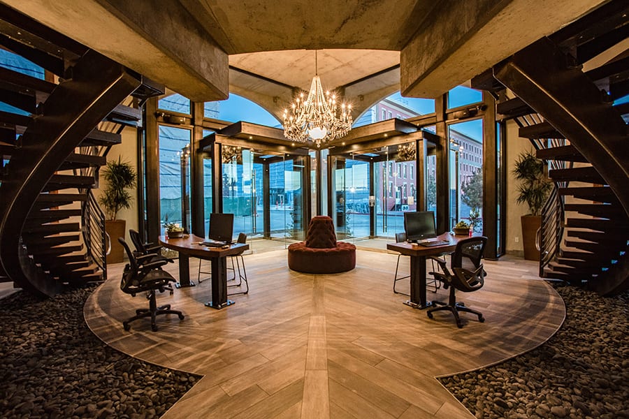 Grand entrance hall with curved dual staircases, wood and stone floors, and glowing glass chandelier.