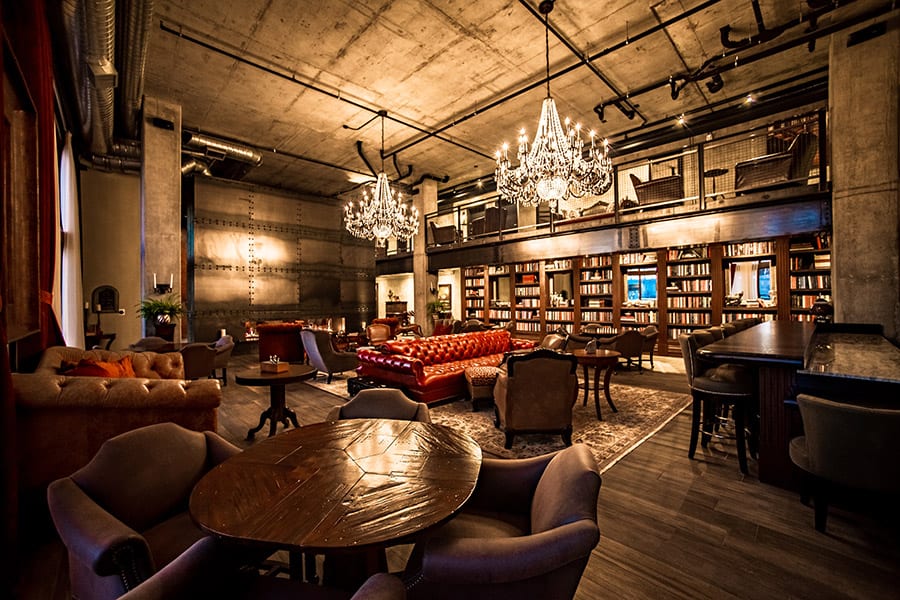 Clubroom with glowing chandeliers, rich couches and chairs, a bookshelf wall, and a circular table.